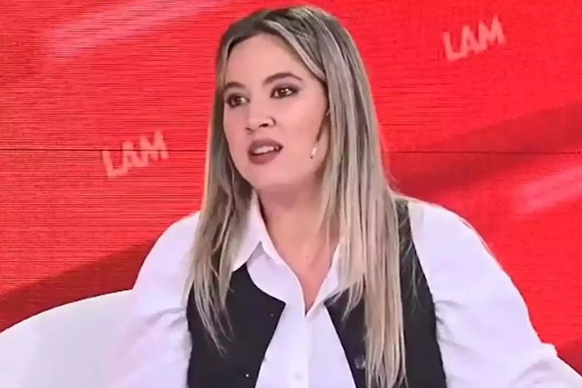 Blonde, straight-haired woman wearing a white jersey and black vest speaking on a television show with a red background and the letters LAM. Blonde, straight-haired woman wearing a white jersey and black vest speaking on a television show with a red background and the letters LAM.