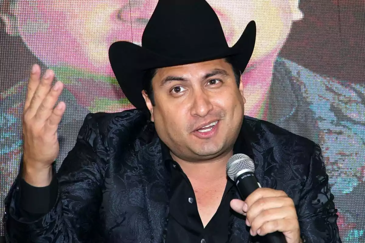 A man with a black hat and decorated jacket holds a microphone while gesturing.