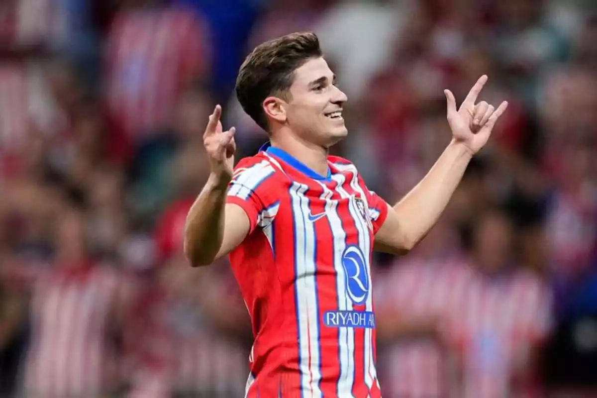 Jugador de fútbol celebrando un gol con los brazos levantados y sonriendo, vistiendo una camiseta a rayas rojas y blancas.