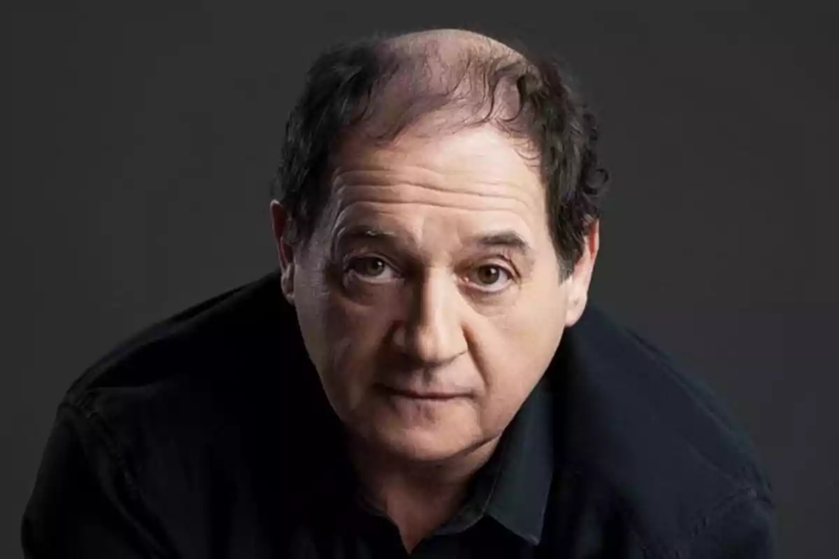 Un hombre de mediana edad con cabello oscuro y escaso mira directamente a la cámara con expresión seria, vistiendo una camisa negra y con un fondo gris oscuro. Un hombre de mediana edad con cabello oscuro y escaso mira directamente a la cámara con expresión seria, vistiendo una camisa negra y con un fondo gris oscuro.