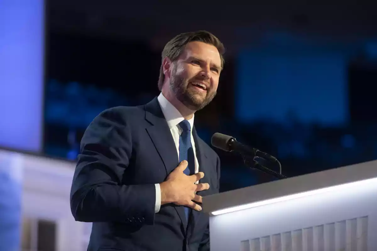 Bearded man in a blue suit smiling while speaking into a microphone on a brightly lit stage