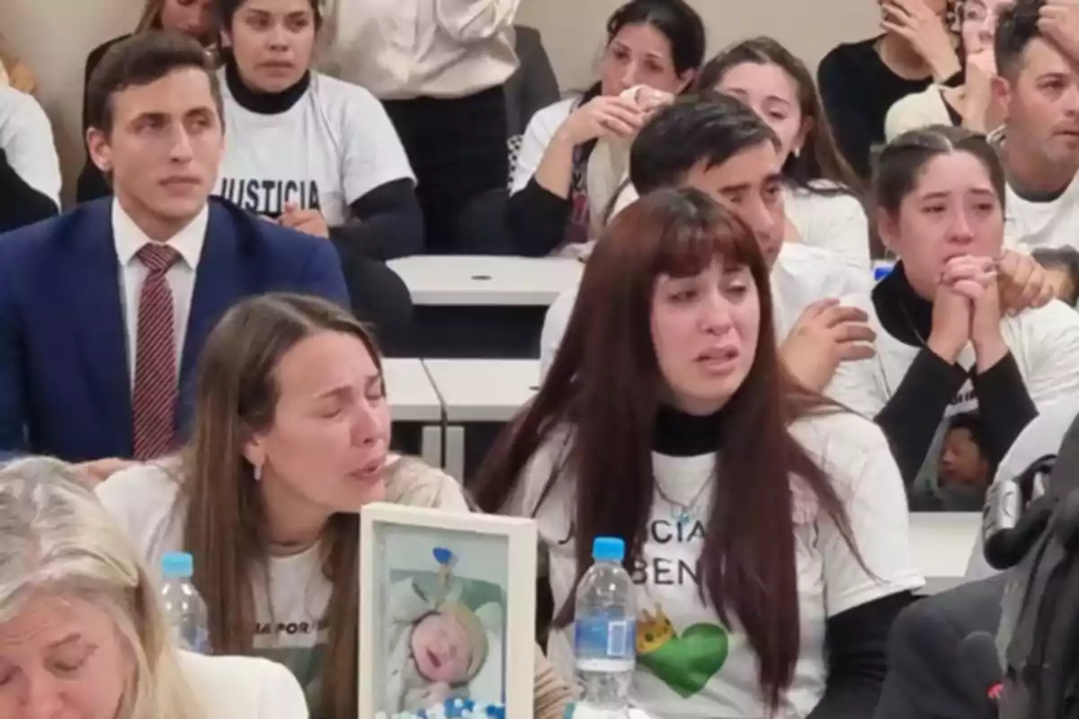 Personas llorando y mostrando dolor en una sala, algunas llevan camisetas blancas con mensajes de justicia y una mujer sostiene una foto de un bebé Personas llorando y mostrando dolor en una sala, algunas llevan camisetas blancas con mensajes de justicia y una mujer sostiene una foto de un bebé