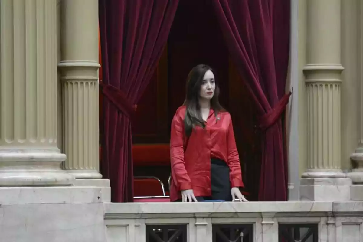 Mujer de pie en un balcón con cortinas rojas y columnas decorativas.