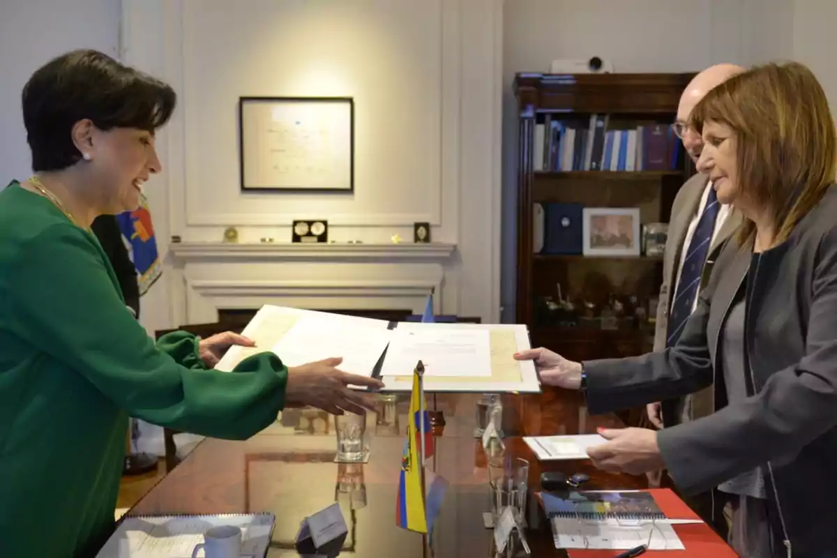 Dos personas intercambiando documentos en una oficina formal, con una bandera sobre la mesa y estantes con libros al fondo.