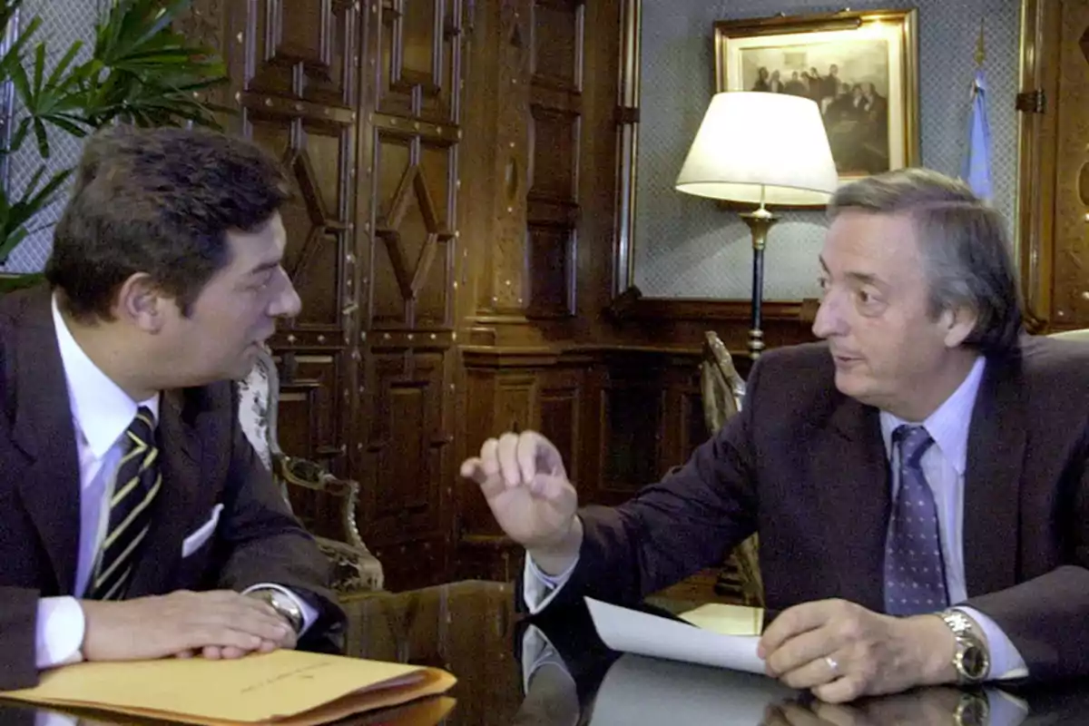Dos hombres de traje conversan sentados en una oficina elegante con paredes de madera y una lámpara encendida al fondo