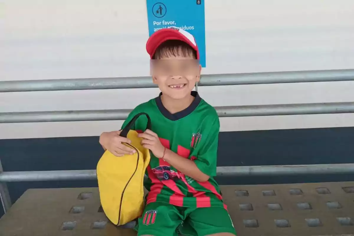 Niño sonriente con uniforme deportivo verde y rojo, gorra roja y mochila amarilla sentado en una banca de metal