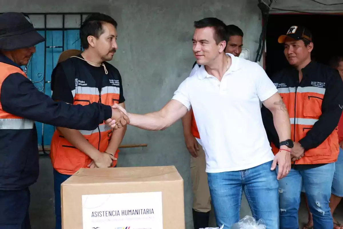 A group of people, some wearing orange vests, is at a humanitarian aid event; two of them are shaking hands in front of a labeled box.