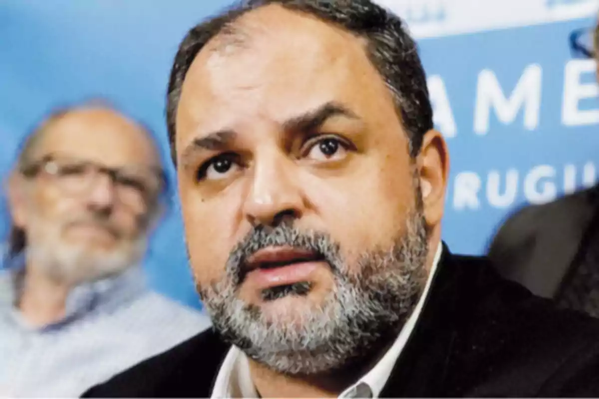 Man with a beard and short hair with a serious expression in the foreground and a blurred blue background with another person in the distance