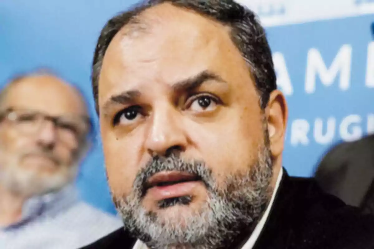 Primer plano de un hombre de barba canosa con expresión seria y fondo azul con letras blancas desenfocadas Primer plano de un hombre de barba canosa con expresión seria y fondo azul con letras blancas desenfocadas
