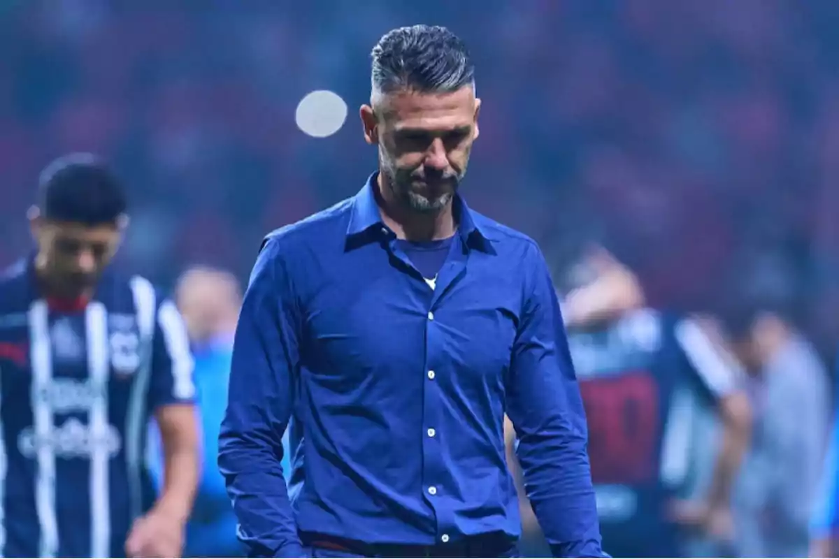 Un hombre con camisa azul camina cabizbajo en un campo de fútbol mientras otras personas están desenfocadas en el fondo.