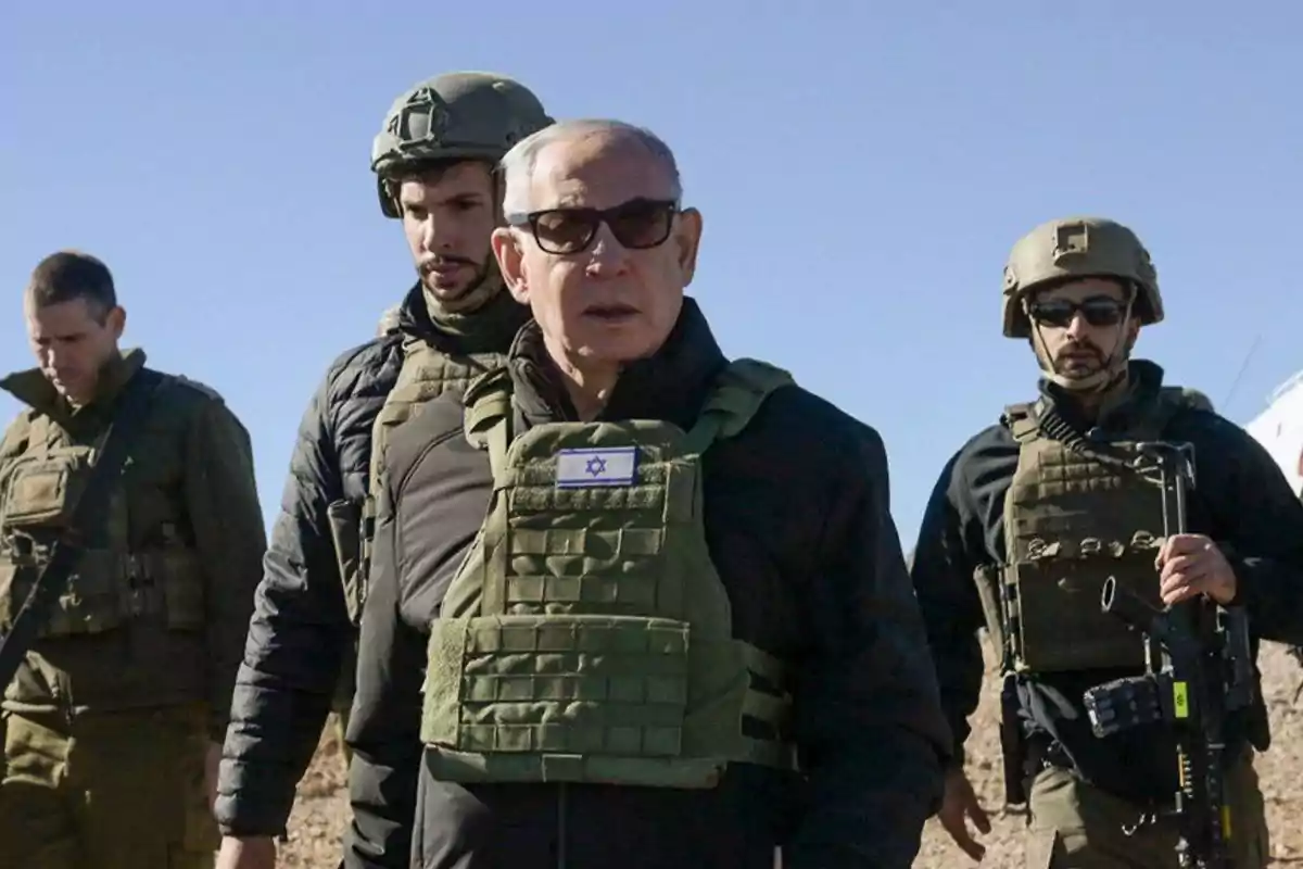A man wearing a bulletproof vest and sunglasses walks accompanied by three soldiers in military uniforms and tactical gear under a clear sky. A man wearing a bulletproof vest and sunglasses walks accompanied by three soldiers in military uniforms and tactical gear under a clear sky.