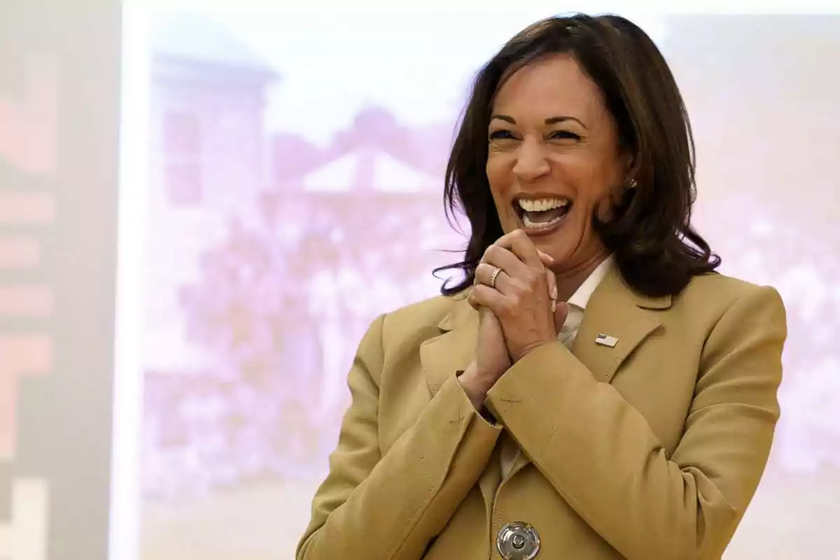 Una mujer sonriente con un traje beige y una insignia en la solapa, con las manos juntas frente a ella.