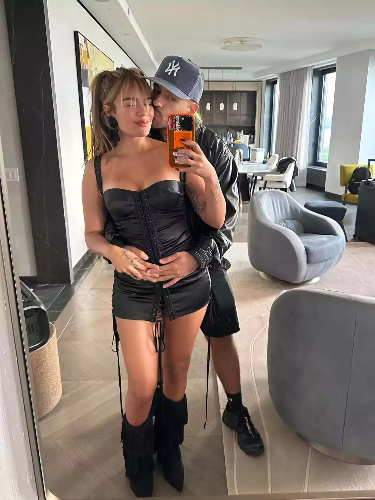 Pareja tomándose una selfie frente a un espejo en una sala moderna, la mujer lleva un vestido negro ajustado y botas con flecos, mientras el hombre la abraza por detrás y lleva gorra gris Pareja tomándose una selfie frente a un espejo en una sala moderna, la mujer lleva un vestido negro ajustado y botas con flecos, mientras el hombre la abraza por detrás y lleva gorra gris