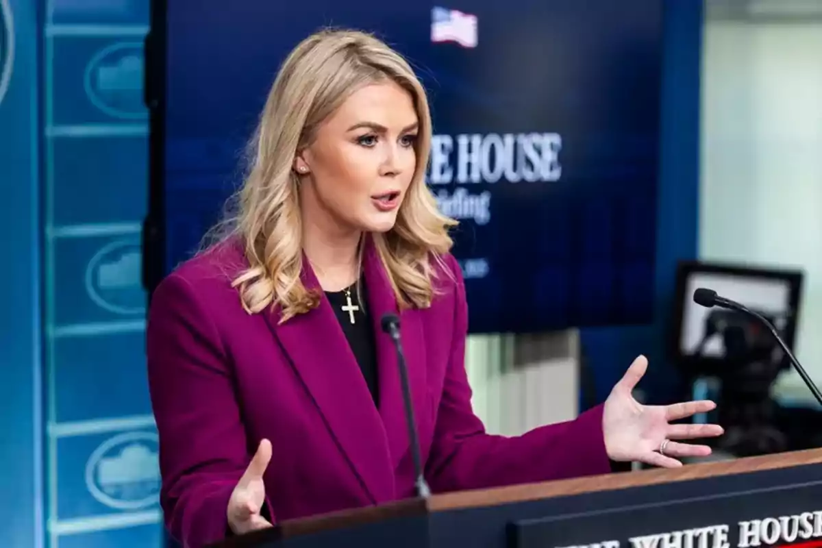 A woman in a purple jacket and a cross necklace speaks at a podium with microphones, with a background showing a White House sign.