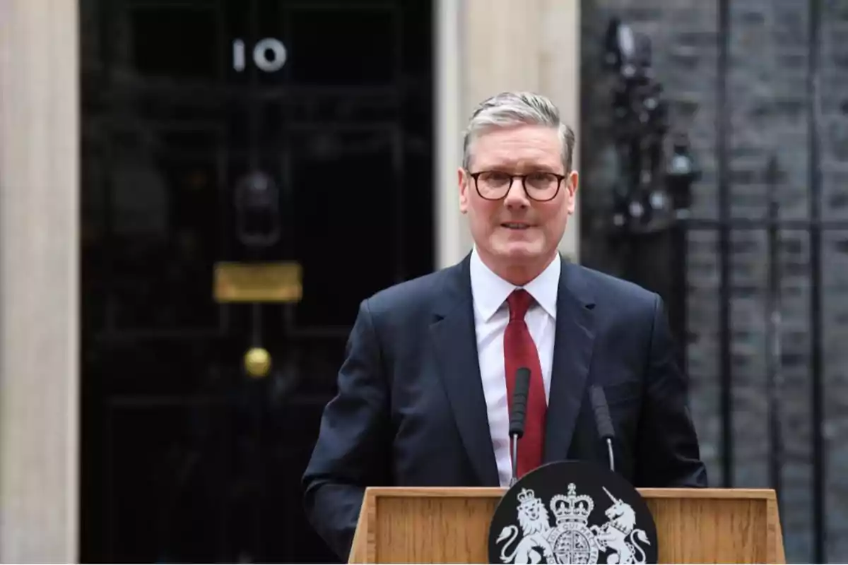 A man with glasses and a dark suit speaks in front of a lectern with microphones and an official emblem on a black door with the number 10. A man with glasses and a dark suit speaks in front of a lectern with microphones and an official emblem on a black door with the number 10.