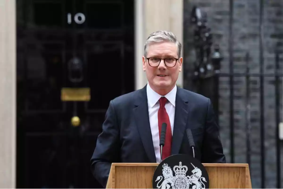 Un hombre con gafas y traje oscuro habla frente a un atril con el emblema del Reino Unido, con la puerta número 10 de Downing Street de fondo.