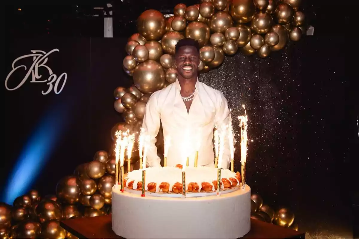 Una persona sonriente está de pie detrás de un gran pastel de cumpleaños con velas encendidas y decoraciones de globos dorados al fondo.