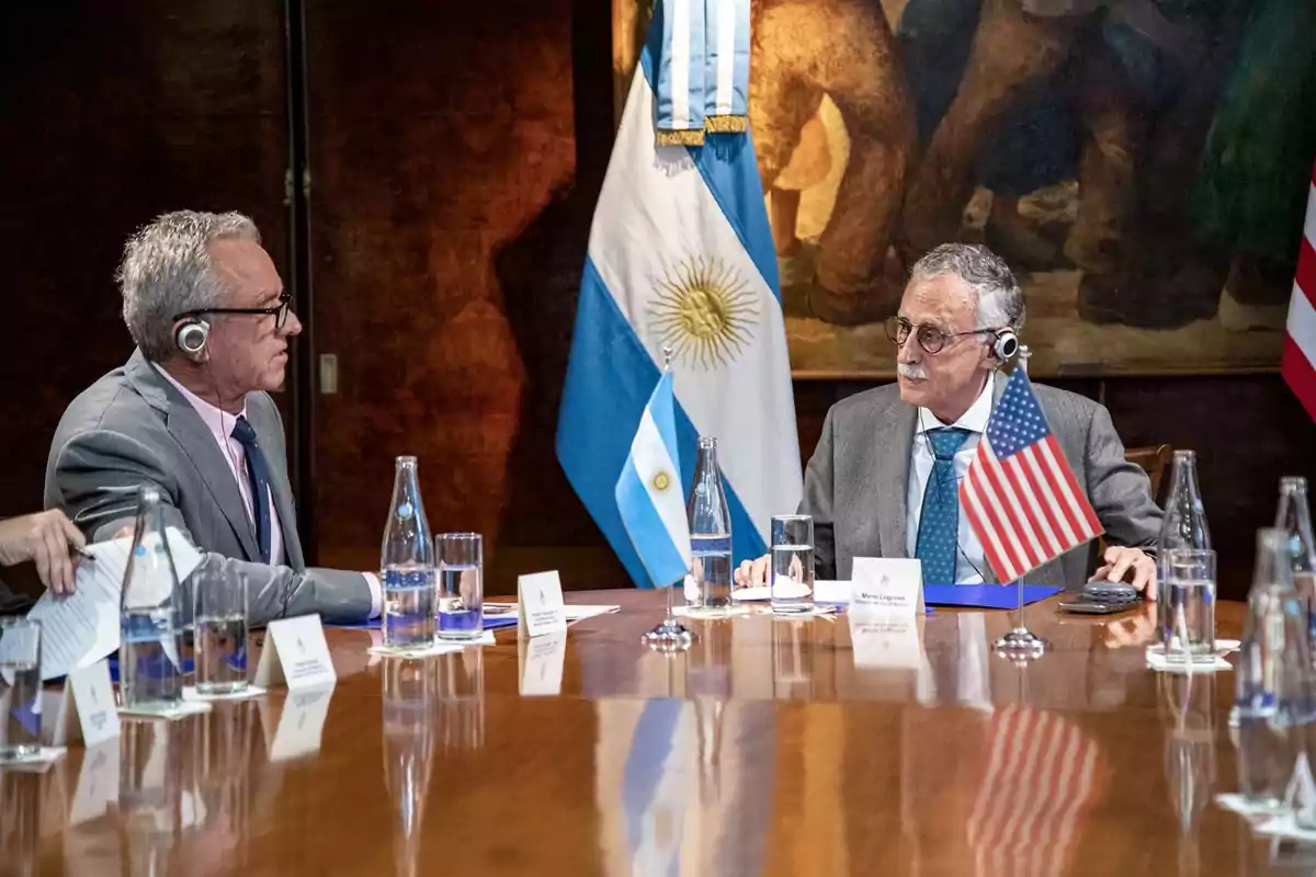 Dos hombres con auriculares de traducción conversan en una mesa de reuniones con banderas de Argentina y Estados Unidos, rodeados de botellas de agua y papeles Dos hombres con auriculares de traducción conversan en una mesa de reuniones con banderas de Argentina y Estados Unidos, rodeados de botellas de agua y papeles