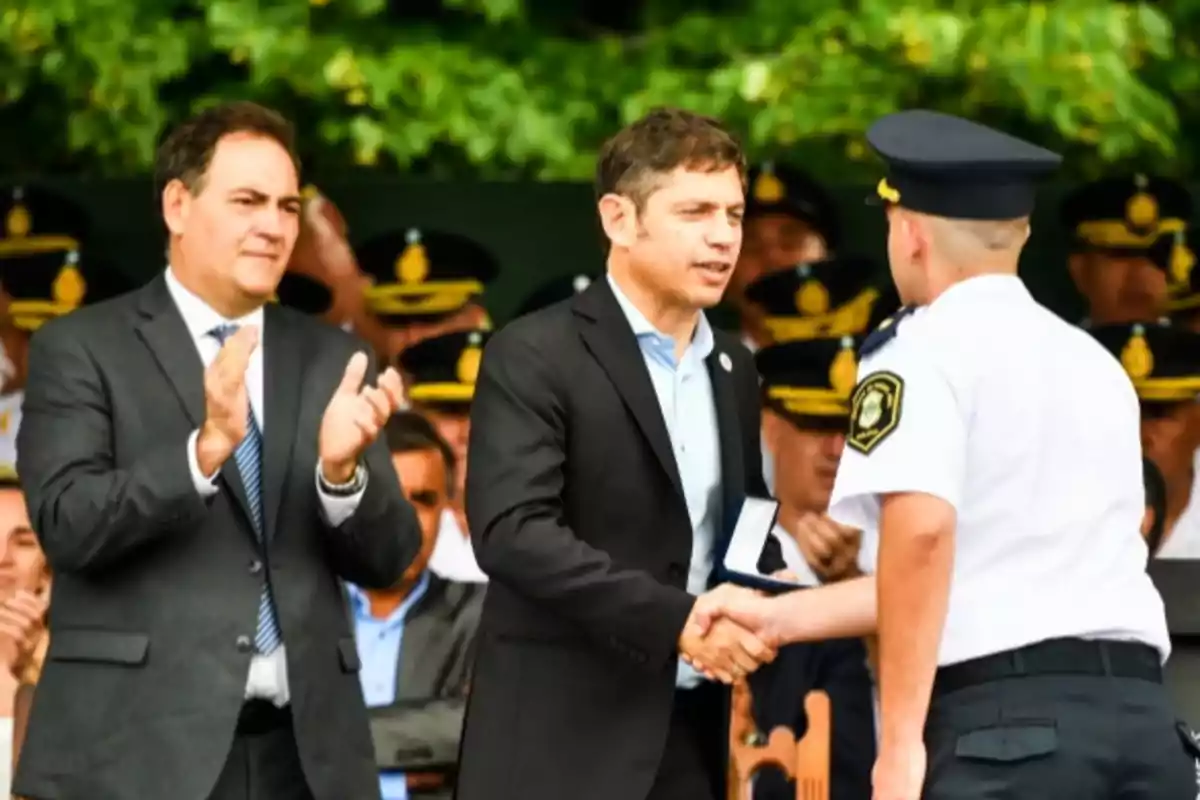 Un hombre de traje aplaude mientras otro hombre de traje entrega un reconocimiento a un oficial de policía en un evento formal al aire libre.