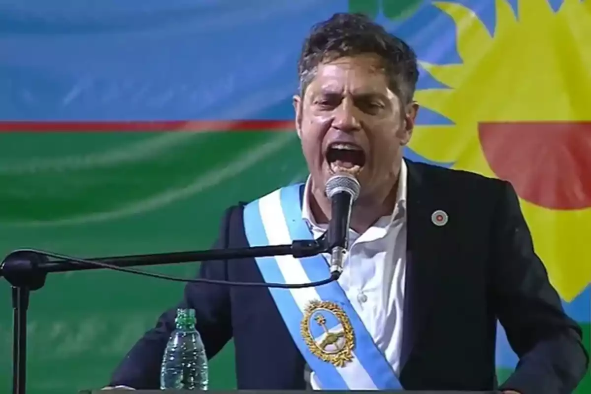 Un hombre con una banda azul y blanca hablando en un micrófono durante un evento, con una bandera colorida de fondo.