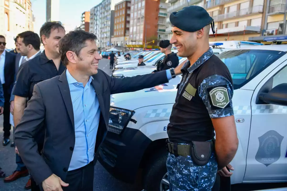 Un hombre de traje sonríe y pone su mano en el hombro de un policía uniformado mientras conversan frente a una fila de vehículos policiales estacionados en una calle urbana.