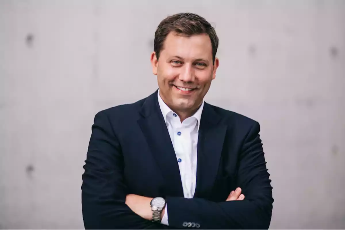 Hombre sonriendo con traje oscuro y camisa blanca, brazos cruzados, fondo gris. Hombre sonriendo con traje oscuro y camisa blanca, brazos cruzados, fondo gris.