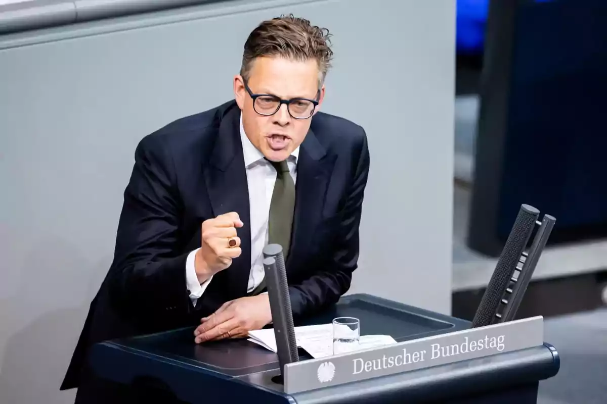Un hombre con gafas y traje oscuro gesticula mientras habla en un podio del Bundestag alemán. Un hombre con gafas y traje oscuro gesticula mientras habla en un podio del Bundestag alemán.