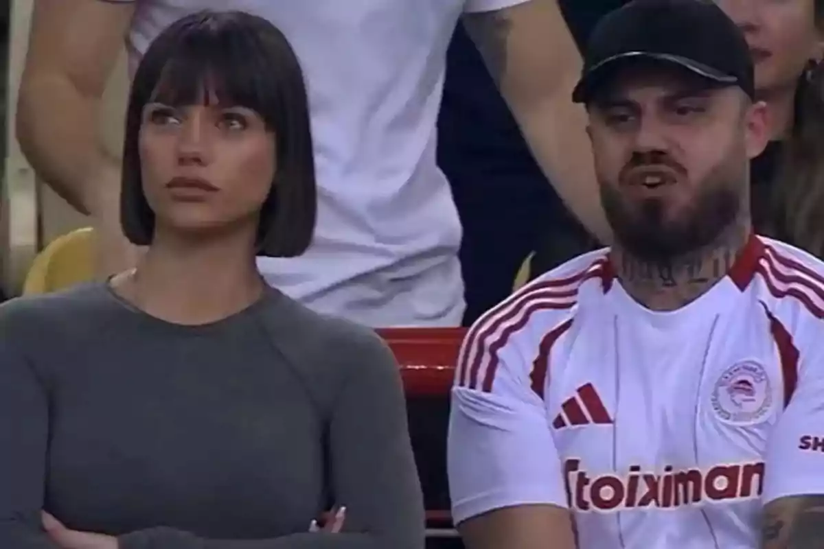 Una mujer y un hombre sentados en un evento deportivo, ella con expresión seria y él con una camiseta deportiva blanca y roja.