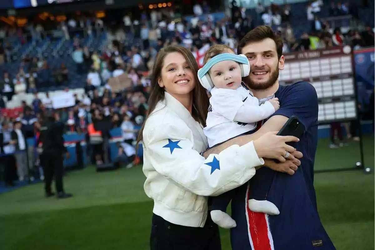 Una pareja sonriente sostiene a un bebé con orejeras en un estadio de fútbol lleno de gente Una pareja sonriente sostiene a un bebé con orejeras en un estadio de fútbol lleno de gente