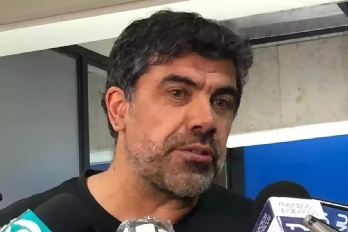 Dark-haired, bearded man speaking in front of several microphones in an interview