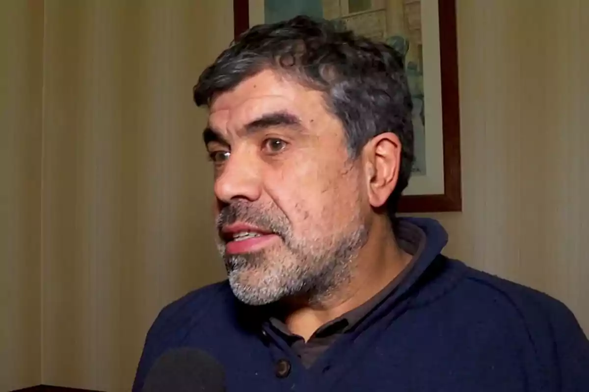 Hombre de cabello canoso y barba hablando frente a un micrófono en una habitación con paredes claras y un cuadro en el fondo Hombre de cabello canoso y barba hablando frente a un micrófono en una habitación con paredes claras y un cuadro en el fondo
