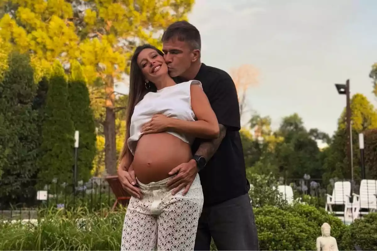 Una pareja sonriente en un jardín con la mujer embarazada mostrando su barriga mientras el hombre la abraza y besa en la mejilla.