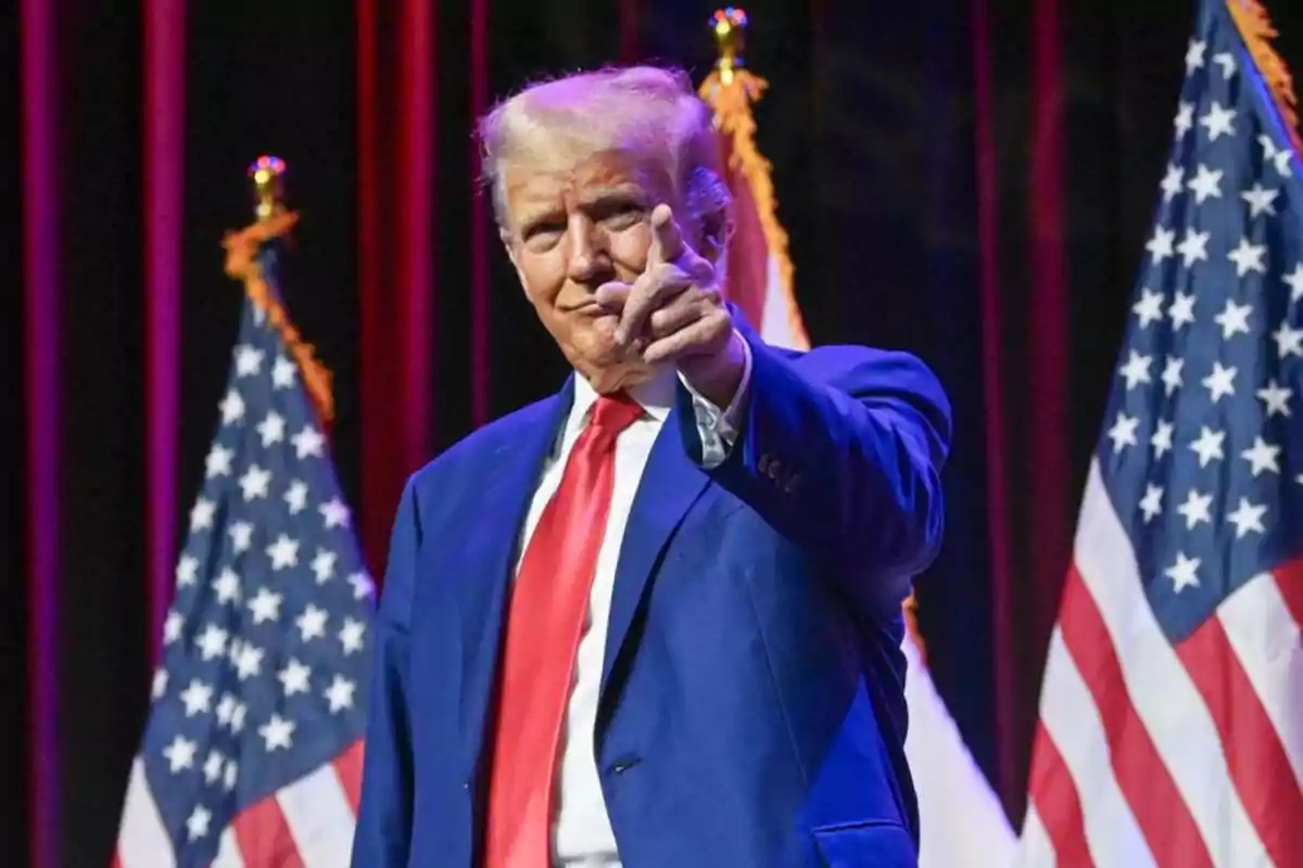 Un hombre de traje azul y corbata roja señala hacia adelante, con banderas de Estados Unidos de fondo.