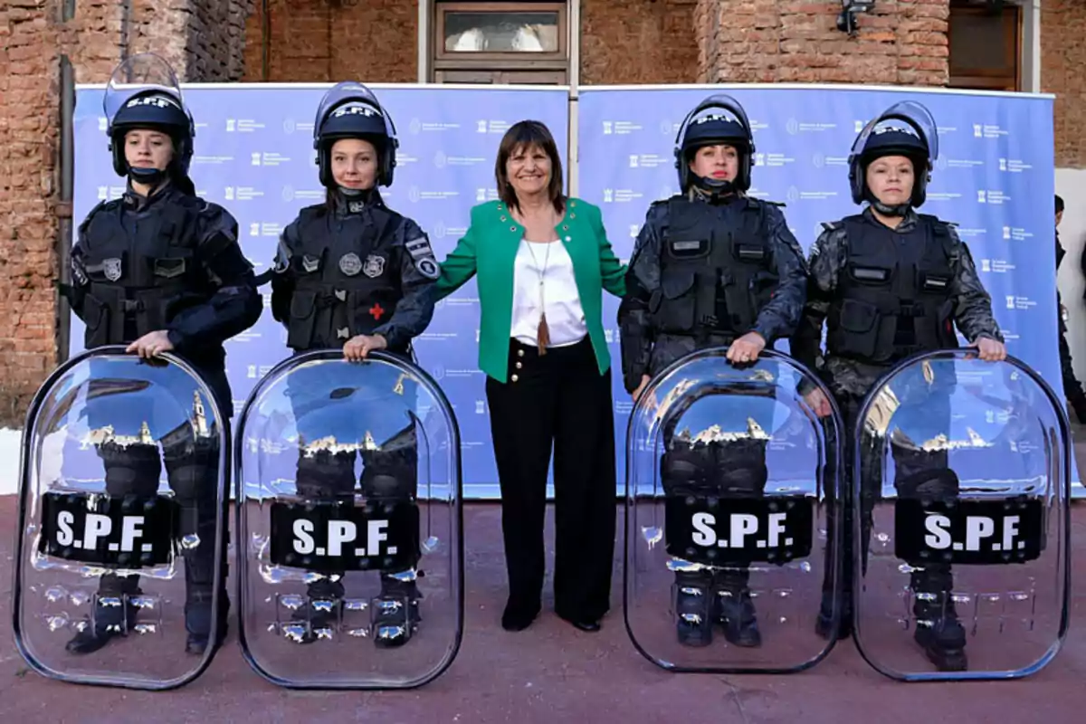 Una mujer vestida de verde está de pie junto a cuatro agentes de policía con equipo antidisturbios y escudos transparentes frente a un fondo azul.