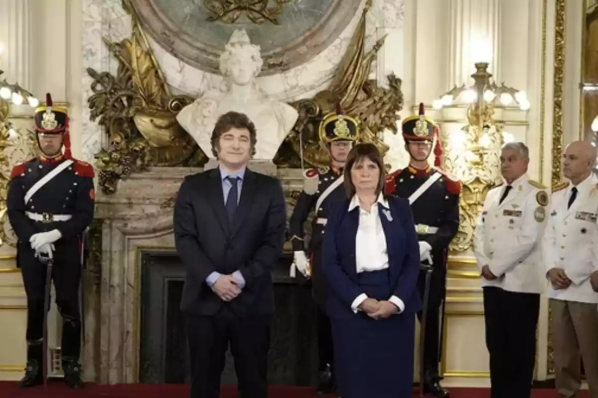 Un grupo de personas posando en un salón elegante con guardias uniformados y una estatua decorativa al fondo.