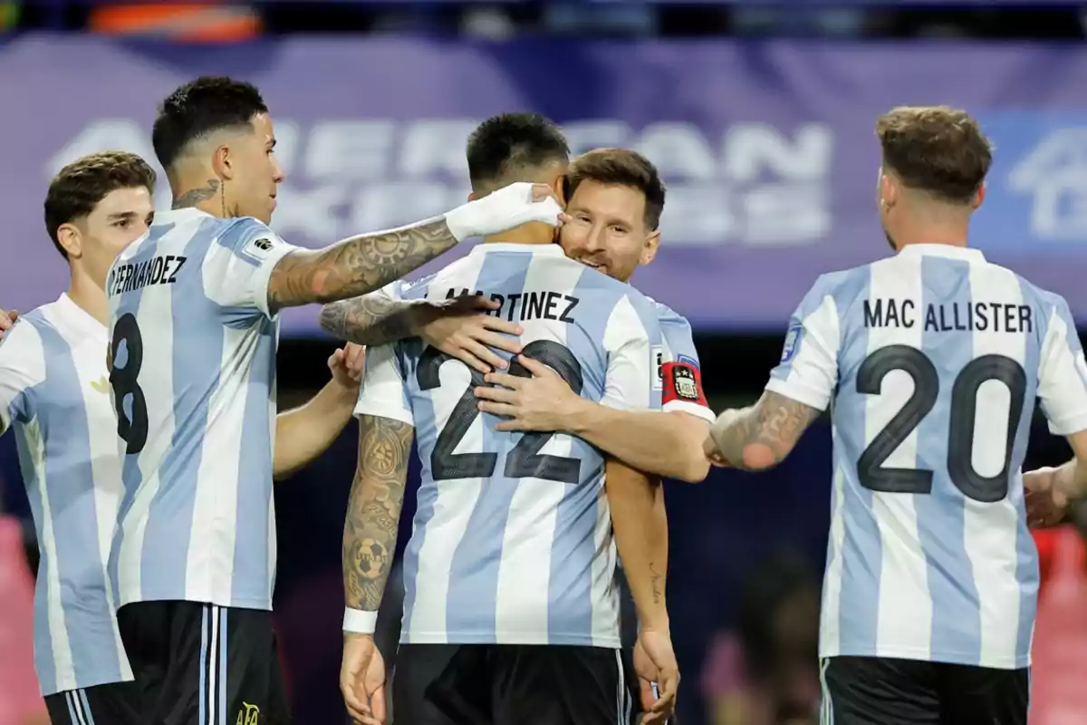 Jugadores de fútbol de Argentina celebran en el campo con abrazos y sonrisas, vistiendo uniformes a rayas celestes y blancas.