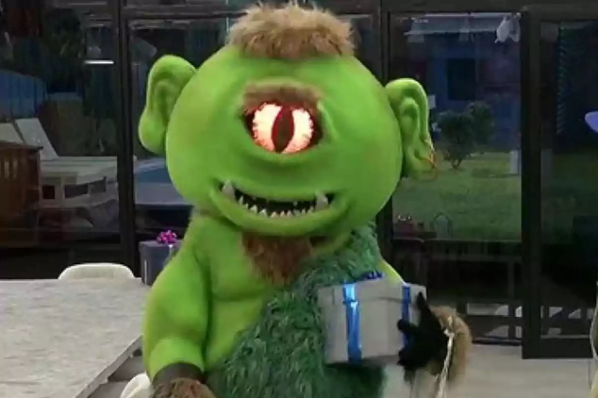 Un personaje disfrazado de cíclope verde con un solo ojo grande y dientes puntiagudos sostiene una caja de regalo plateada con listón azul en un entorno interior. Un personaje disfrazado de cíclope verde con un solo ojo grande y dientes puntiagudos sostiene una caja de regalo plateada con listón azul en un entorno interior.