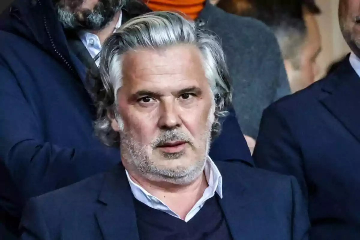 A gray-haired man with a short beard wears a dark suit and a white jersey while looking ahead in a busy environment. A gray-haired man with a short beard wears a dark suit and a white jersey while looking ahead in a busy environment.