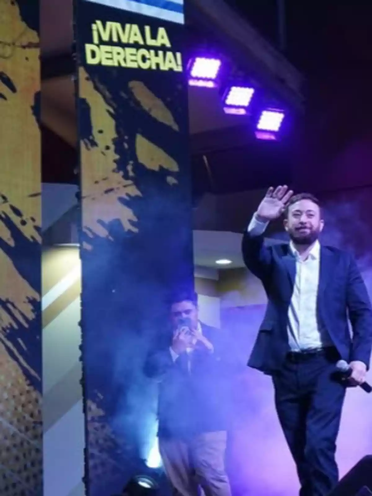 A man in a suit greets the audience while holding a microphone, with a sign that says A man in a suit greets the audience while holding a microphone, with a sign that says