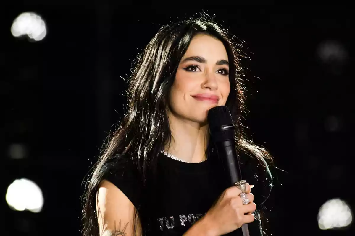 Mujer de cabello largo y oscuro sonríe mientras sostiene un micrófono en un escenario iluminado