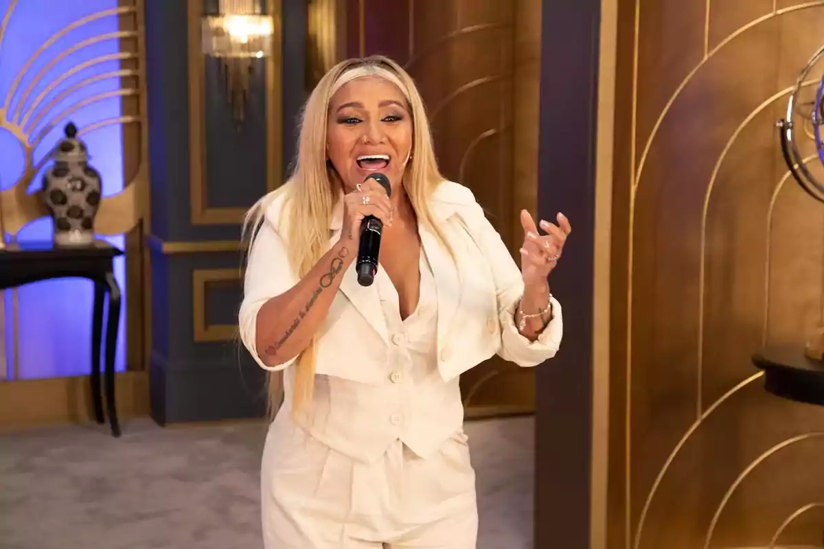 Una mujer con cabello largo y rubio está cantando con un micrófono en la mano, vestida con un conjunto blanco elegante, en un ambiente decorado con detalles dorados y azules.