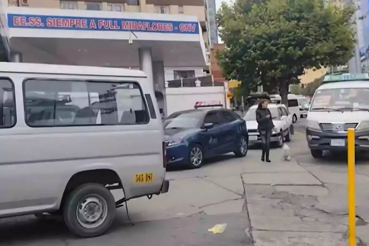 Una calle transitada con varios vehículos estacionados y una persona caminando con un perro frente a una estación de servicio.