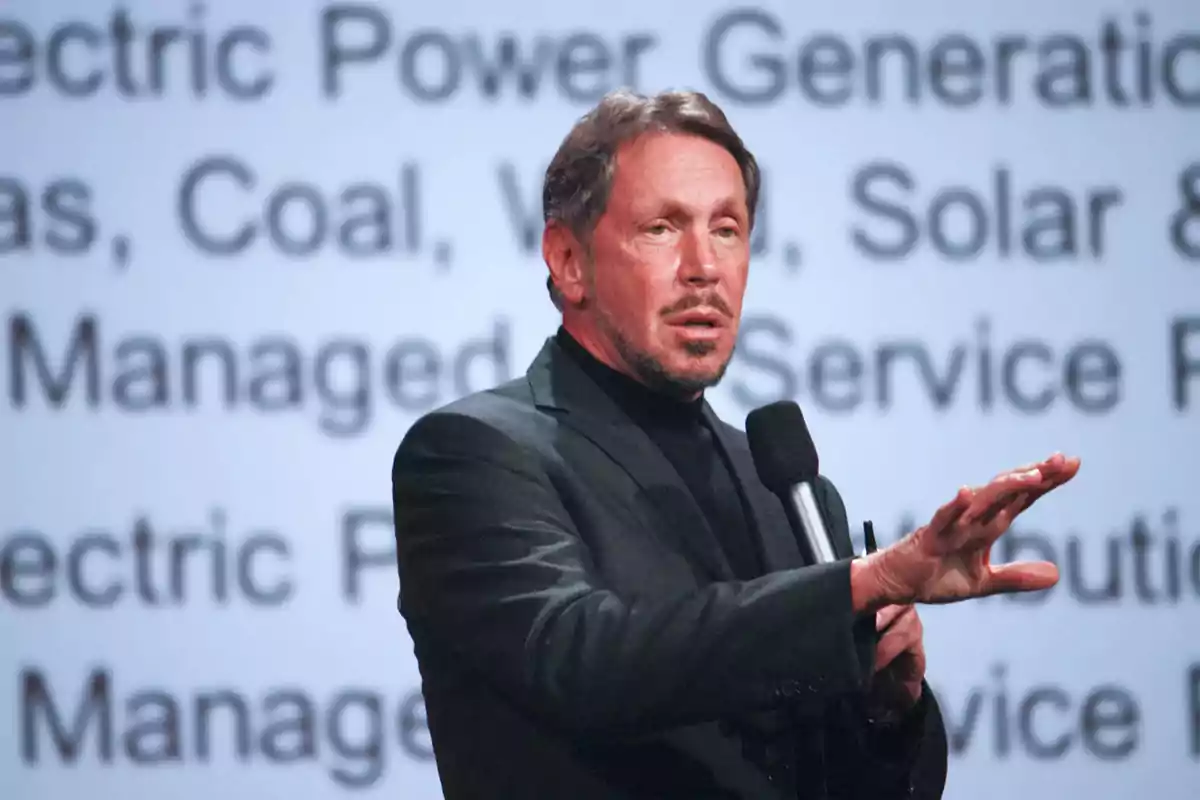 Middle-aged man with a beard and short hair wearing a dark jacket while holding a microphone and gesturing with his hand in front of a screen with blurred text in the background