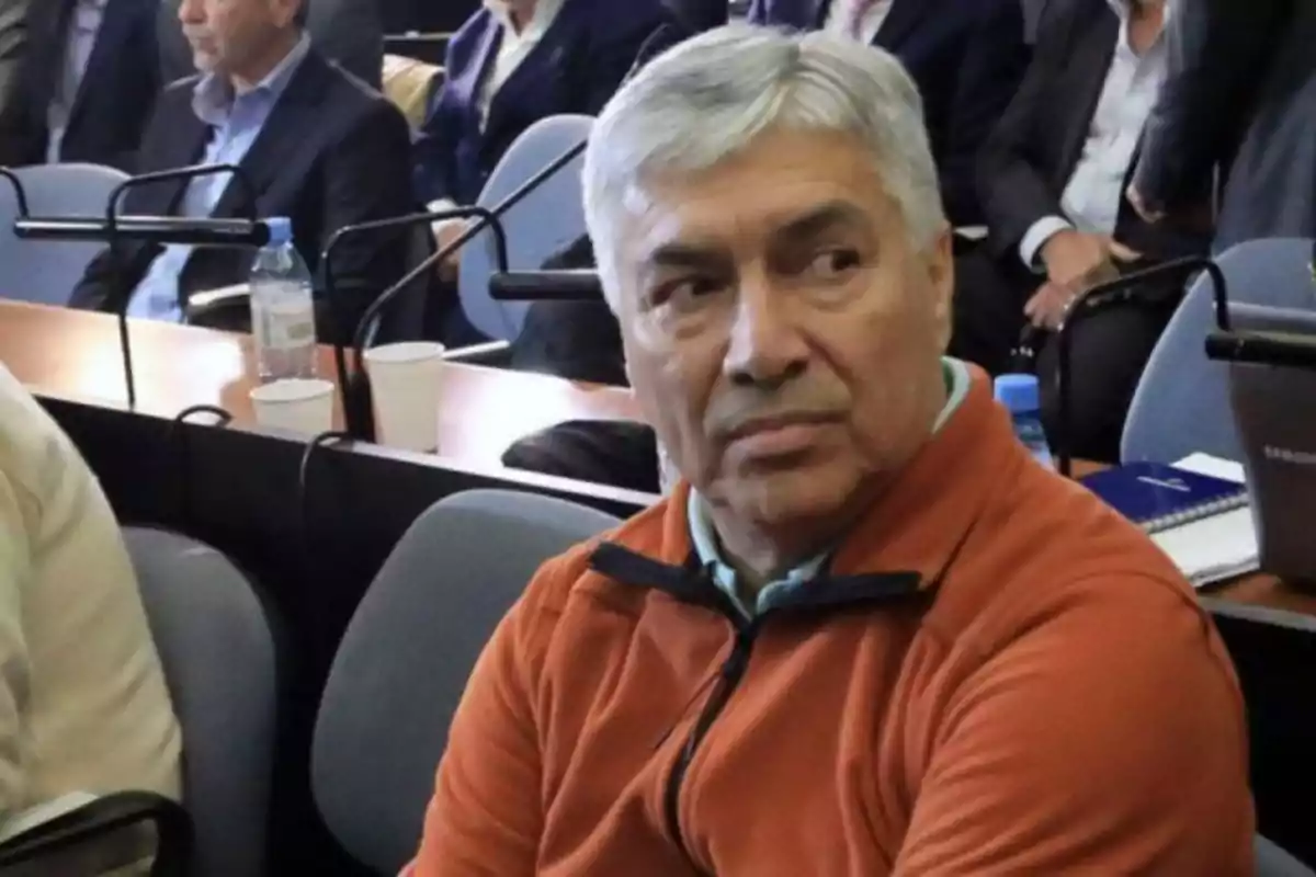 Un hombre de cabello canoso y suéter naranja está sentado en una sala de conferencias rodeado de otras personas.