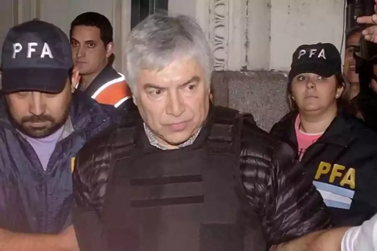 Hombre canoso escoltado por agentes de la policía federal argentina con chalecos y gorras que llevan las siglas PFA Hombre canoso escoltado por agentes de la policía federal argentina con chalecos y gorras que llevan las siglas PFA
