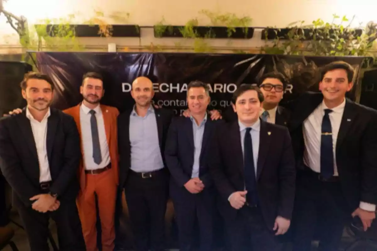 A group of men in suits posing together in front of a dark background with decorative plants at the top.