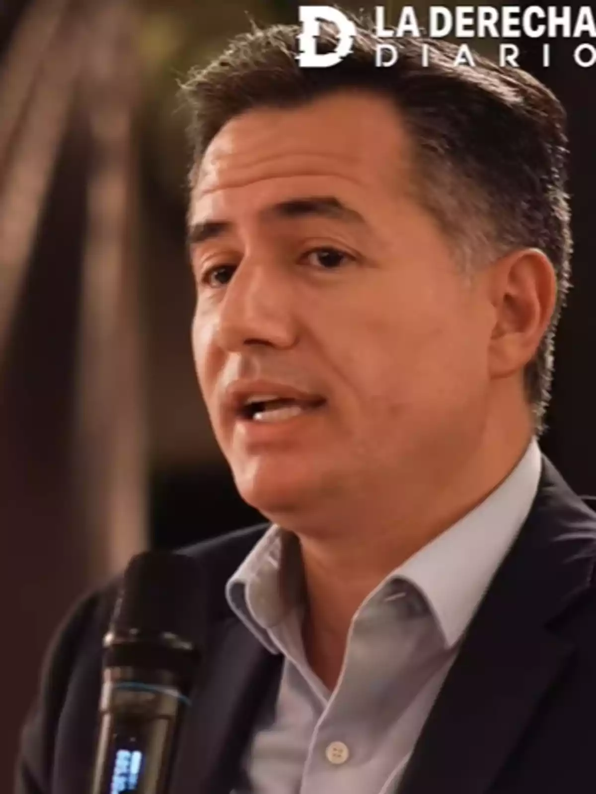A man speaking into a microphone with a blurred background and a logo in the top right corner. A man speaking into a microphone with a blurred background and a logo in the top right corner.