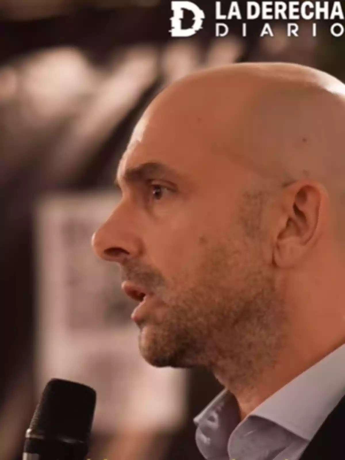A bald man speaking into a microphone with a blurred background and a logo in the top right corner. A bald man speaking into a microphone with a blurred background and a logo in the top right corner.