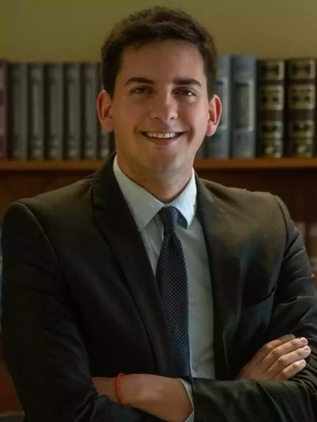 A young man in a suit and tie smiles with his arms crossed in front of a bookshelf. A young man in a suit and tie smiles with his arms crossed in front of a bookshelf.