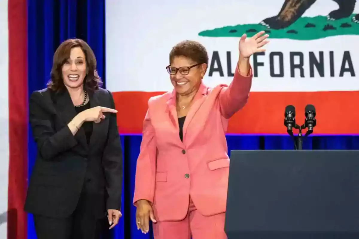 Two women smile on a stage in front of a California flag; one waves with her hand raised while the other points at her, and both are wearing formal suits. Two women smile on a stage in front of a California flag; one waves with her hand raised while the other points at her, and both are wearing formal suits.